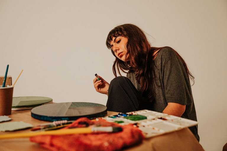 A thoughtful female artist holds a paint bottle while surrounded by her tools in an art studio.